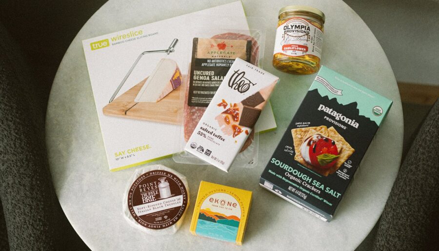 A round table displays a cheese slicer, uncured salami, sea salt crispbread, a jar of marinated olives, chocolate, smoked salmon, and a round tin of cheese, all in their packaging.