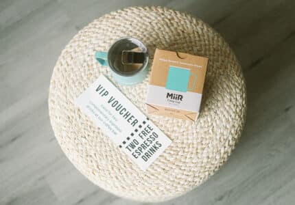A teal camp cup with tea, a box labeled "MiiR Camp Cup," and a VIP voucher for two free espresso drinks rest on a round woven ottoman, viewed from above.