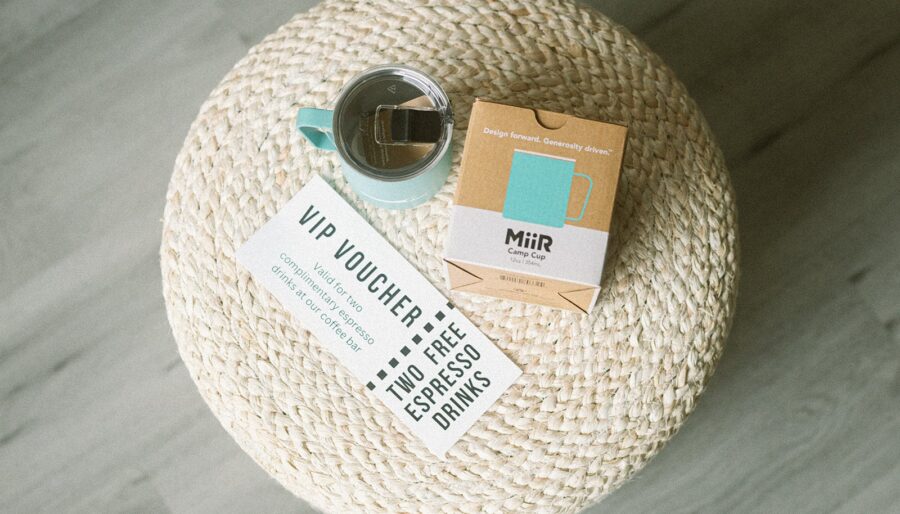 A teal camp cup with tea, a box labeled "MiiR Camp Cup," and a VIP voucher for two free espresso drinks rest on a round woven ottoman, viewed from above.