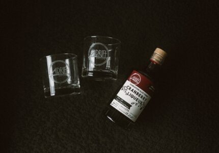 A bottle of cranberry liqueur with a wooden cap lies on a dark surface next to two empty clear glasses, both labeled with the Drift Distillery logo.