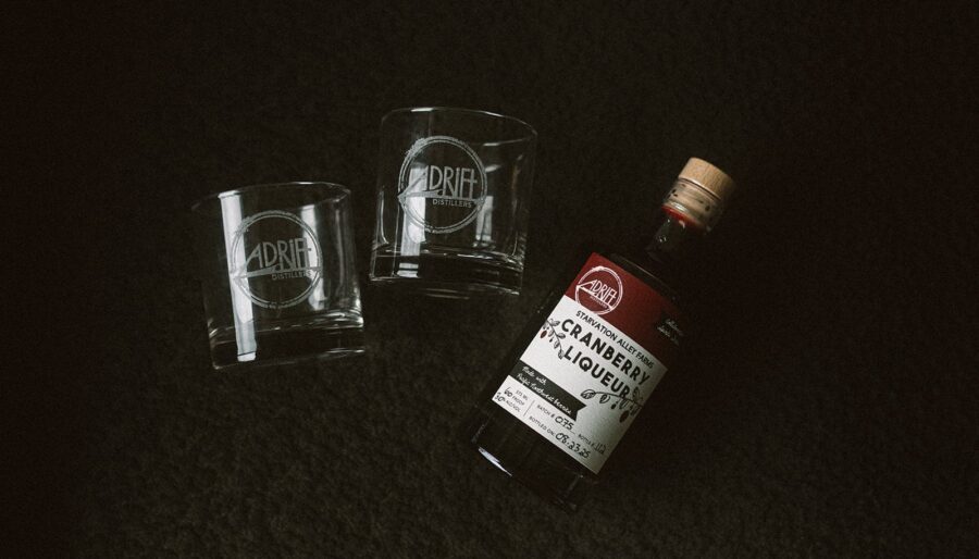 A bottle of cranberry liqueur with a wooden cap lies on a dark surface next to two empty clear glasses, both labeled with the Drift Distillery logo.