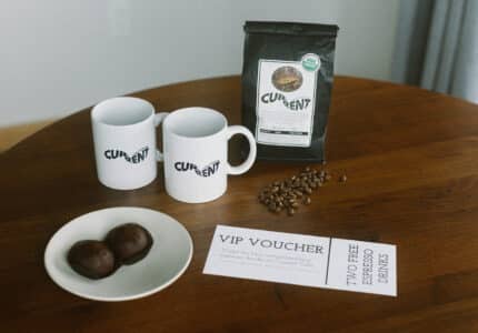 Two white mugs and a black bag of coffee labeled "Current" are on a wooden table, along with coffee beans, a plate with two chocolate truffles, and a VIP voucher for two free espresso drinks.