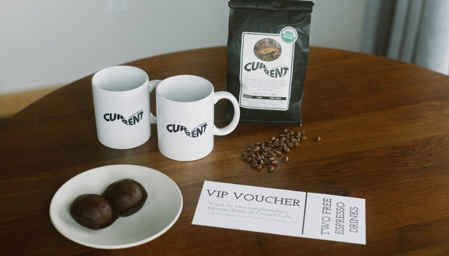 Two white mugs and a black bag of coffee labeled "Current" are on a wooden table, along with coffee beans, a plate with two chocolate truffles, and a VIP voucher for two free espresso drinks.