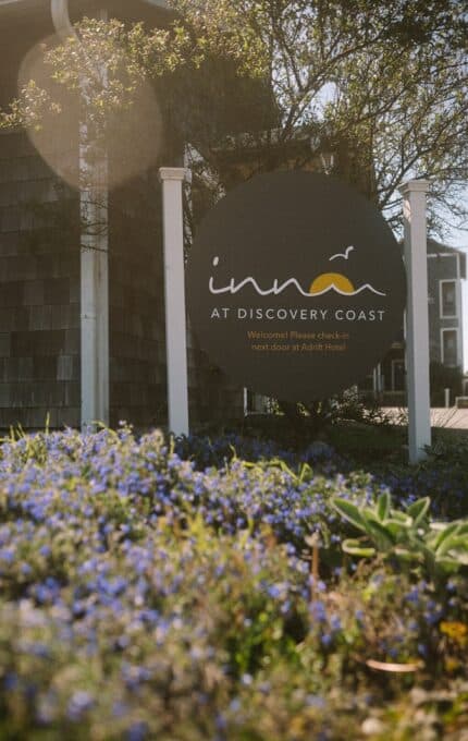 A circular sign for "Inn at Discovery Coast" stands in front of a building, surrounded by green plants and small purple flowers. Sunlight streams through the trees, and text on the sign instructs guests to check in next door.