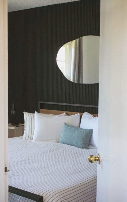 A neatly made bed with white and striped bedding is against a dark textured wall. A modern, irregularly shaped mirror hangs above the bed. A window is reflected in the mirror, and the door is partially open.
