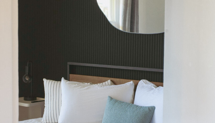 A neatly made bed with white and striped bedding is against a dark textured wall. A modern, irregularly shaped mirror hangs above the bed. A window is reflected in the mirror, and the door is partially open.