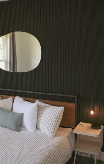 A modern bedroom with a dark accent wall, a round mirror, a framed sunset artwork, a bed with white striped bedding, and two small white nightstands with hanging lamps on either side.