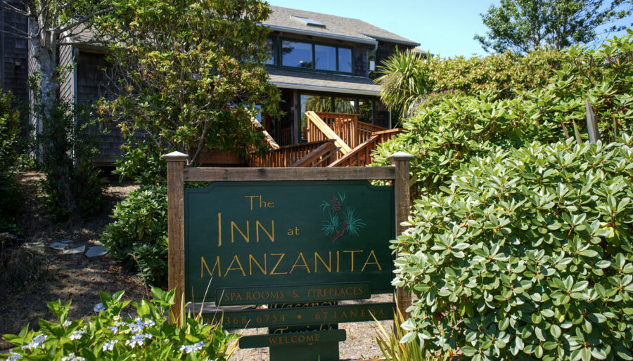 A green sign reading “The Inn at Manzanita” stands among lush bushes and trees, with a wooden building and stairs visible in the background on a sunny day.