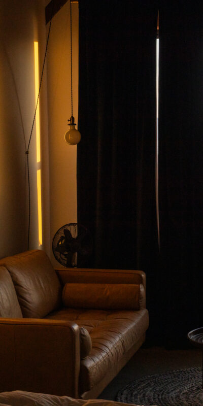 A cozy room with a brown leather sofa, round coffee table, bed corner, and wall art. Warm lighting illuminates the space. Large windows and a door reveal a view of ships on the water outside.