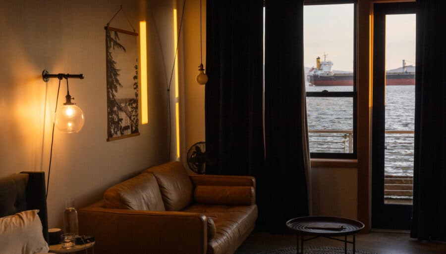 A cozy room with a brown leather sofa, round coffee table, bed corner, and wall art. Warm lighting illuminates the space. Large windows and a door reveal a view of ships on the water outside.