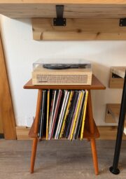 A record player with a clear dust cover sits on top of a small, mid-century style wooden table. Vinyl records are stored vertically on the lower shelf. Wooden shelves and flooring are visible in the background.