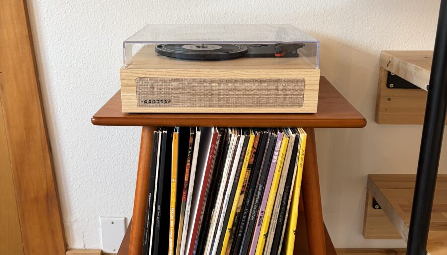 A record player with a clear dust cover sits on top of a small, mid-century style wooden table. Vinyl records are stored vertically on the lower shelf. Wooden shelves and flooring are visible in the background.