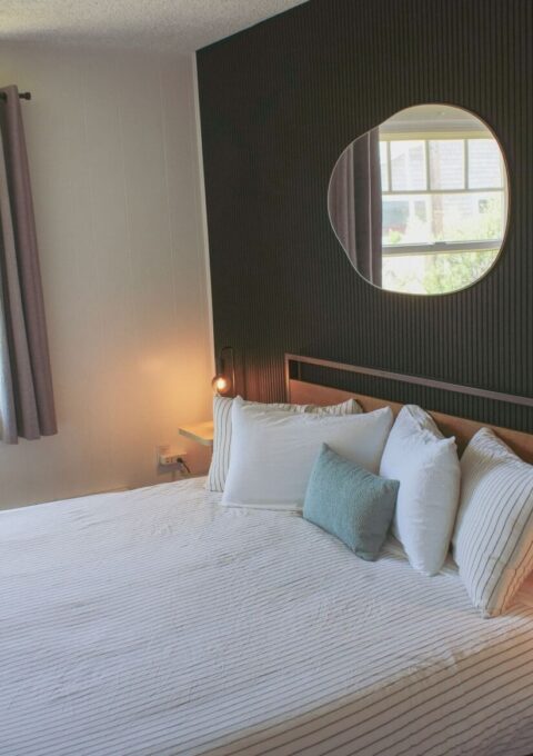 A neatly made bed with white and gray striped bedding sits against a black accent wall, which features a round mirror. Two nightstands with small lamps flank the bed, and sunlight enters through windows with gray curtains.
