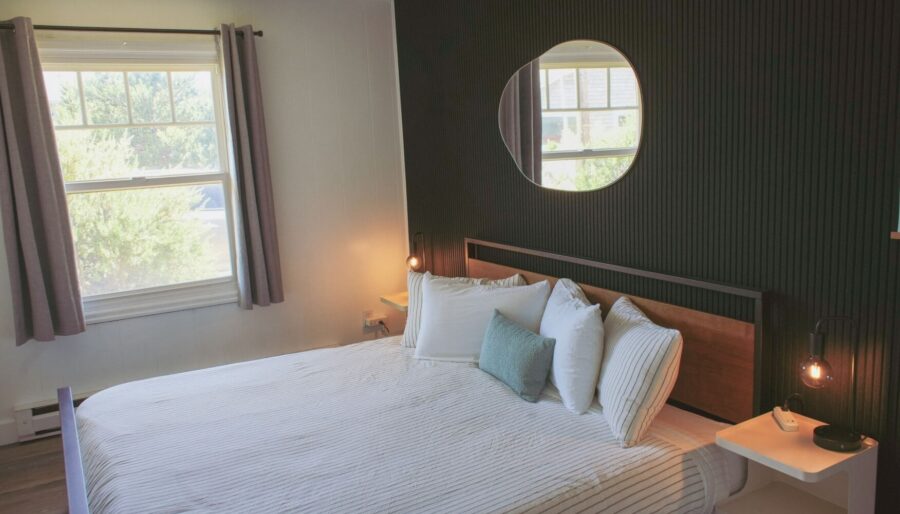 A neatly made bed with white and gray striped bedding sits against a black accent wall, which features a round mirror. Two nightstands with small lamps flank the bed, and sunlight enters through windows with gray curtains.