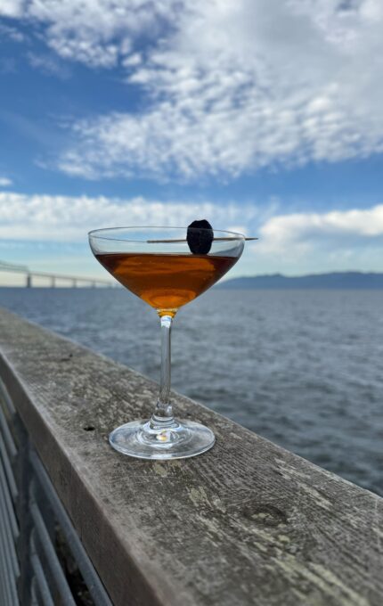 A cocktail in a coupe glass garnished with a dark cherry on a skewer sits on a wooden railing by the water, with a bridge and cloudy sky in the background.