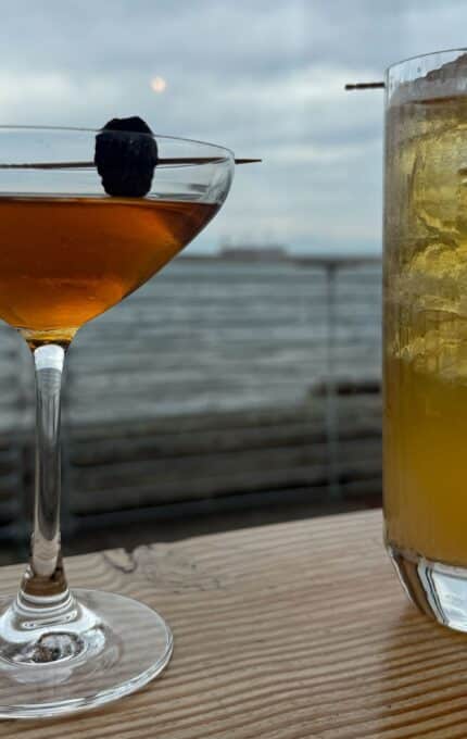 A cocktail in a stemmed glass with a dark garnish on a stick sits next to a tall glass of iced yellow drink, also garnished, on a wooden table with a cloudy waterfront view in the background.