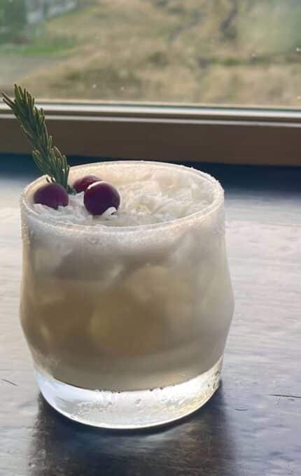 A short glass with a white, iced drink garnished with two berries and a sprig of rosemary sits on a dark wooden surface in front of a window. The rim of the glass is coated with sugar.
