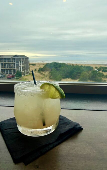 A salt-rimmed glass of a light-colored drink with a lime wedge and straw sits on a black napkin by a window overlooking a coastal landscape with grass, buildings, and the ocean under a cloudy sky.