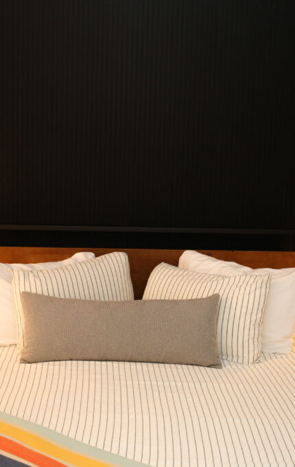 A neatly made bed with striped bedding, a long gray cushion, and several pillows is centered between two small nightstands, each with a lit lamp. The wall behind the bed is black, and a striped blanket is folded at the foot.