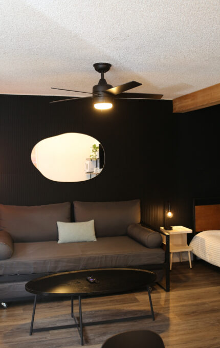A modern living space with a gray sofa, small white side table, bed, black coffee table, wall mirror, and ceiling fan with light, all set against a black accent wall and wood flooring.