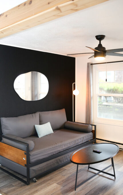 A modern living room with a dark accent wall, oval mirror, gray daybed with cushions, black oval coffee table, floor lamp, ceiling fan, and large window with gray curtains. A remote is on the table.