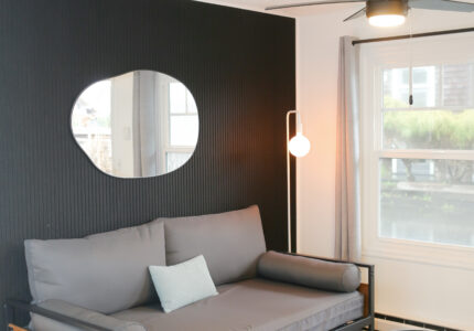 A modern living room with a gray daybed, a small oval black coffee table, a floor lamp with a round bulb, a wall mirror, and a window with gray curtains. The wall behind the daybed is black.