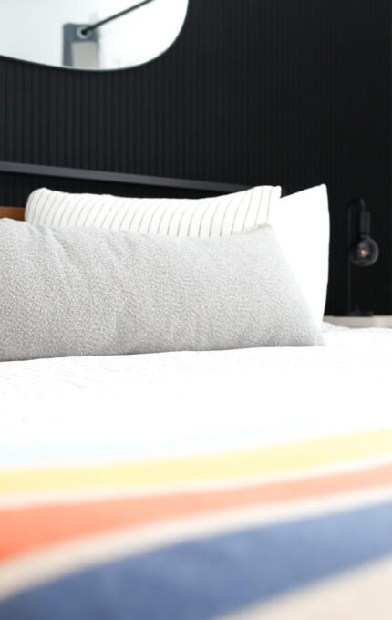 A neatly made bed with a white and striped bedspread, a long gray pillow, and two striped pillows. The bed is against a black paneled wall with a mirror above it. A lamp is visible on a nightstand.