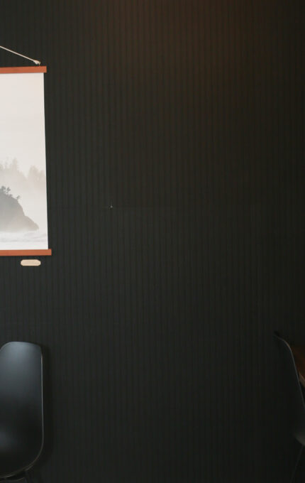 A small corner with a wooden round table and black chair, a black wall with a hanging landscape print, and a shelving unit holding a plant, microwave, mugs, a small fridge, and other kitchen items.