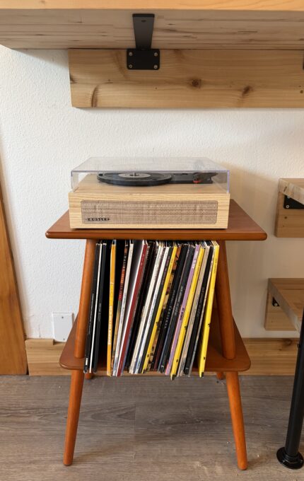 A wooden turntable with a clear dust cover sits on top of a mid-century modern stand, which holds a collection of vinyl records neatly arranged below. The stand is positioned against a wall with wood shelves above it.
