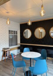 A modern room with blue chairs around a round white table, three round mirrors on a black accent wall, hanging light bulbs, a small bookshelf with books, and a window with a white brick frame.