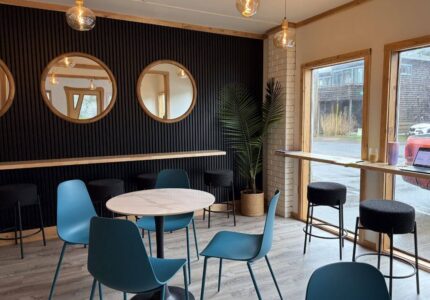 A modern café interior with blue chairs around a white table, high stools at a counter by large windows, three round mirrors on a black accent wall, and a potted plant in the corner. Natural light comes through the windows.