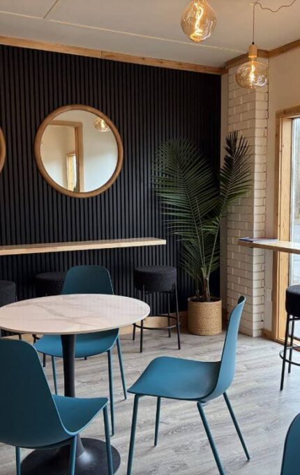 A modern café interior with blue chairs around a white table, high stools at a counter by large windows, three round mirrors on a black accent wall, and a potted plant in the corner. Natural light comes through the windows.