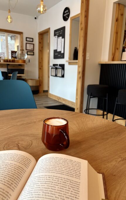 A brown ceramic cup of coffee sits on a wooden table next to an open book in a modern café with wooden accents, blue chairs, and a menu on the wall in the background.
