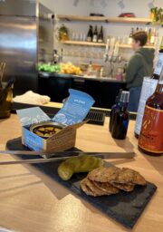 A slate serving board holds a tin of mussels in curry sauce, several round crackers, and sliced pickles on a wooden counter; a person stands behind the bar with bottles, glasses, and fruit visible in the background.