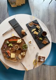 A round white table with two blue chairs holds wooden and slate boards with cheese, meats, bread, olives, nuts, pickles, and small jars. Used utensils and crumbs are scattered on the table.