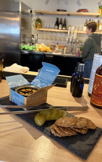 A slate serving board holds a tin of mussels in curry sauce, several round crackers, and sliced pickles on a wooden counter; a person stands behind the bar with bottles, glasses, and fruit visible in the background.