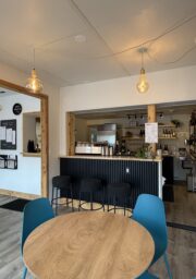 A small café interior with a wooden table and blue chairs in the foreground, a counter with black stools, shelves with bottles and jars, hanging lights, and a menu on the wall.