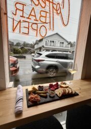 A wooden counter by a window displays a slate with cheeses, meats, bread, and olives, a rolled napkin, and a glass of white wine. Outside, parked cars and houses are visible through a rainy window with neon signage.