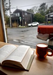 A book lies open on a wooden table next to a cup of coffee in an orange mug by a window. Outside, it's rainy, and houses, parked cars, and wet streets are visible through the window.