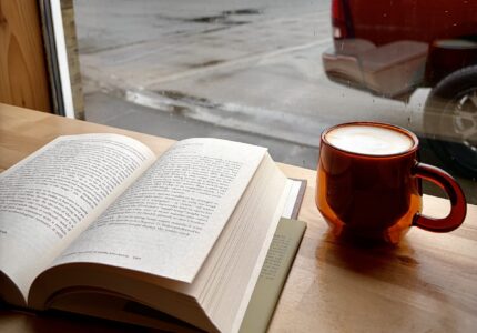 A book lies open on a wooden table next to a cup of coffee in an orange mug by a window. Outside, it's rainy, and houses, parked cars, and wet streets are visible through the window.
