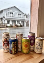 Six colorful cans of craft beer and cider are arranged side by side on a wooden table next to a window, with a rainy street and a white two-story building visible outside.