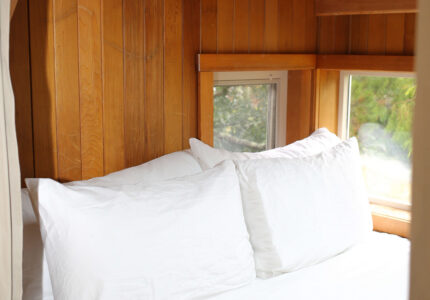 A small cozy bed with white pillows and bedding sits against wooden paneled walls under a sloped ceiling; a wall-mounted light fixture is above the headboard, and windows let in natural light.