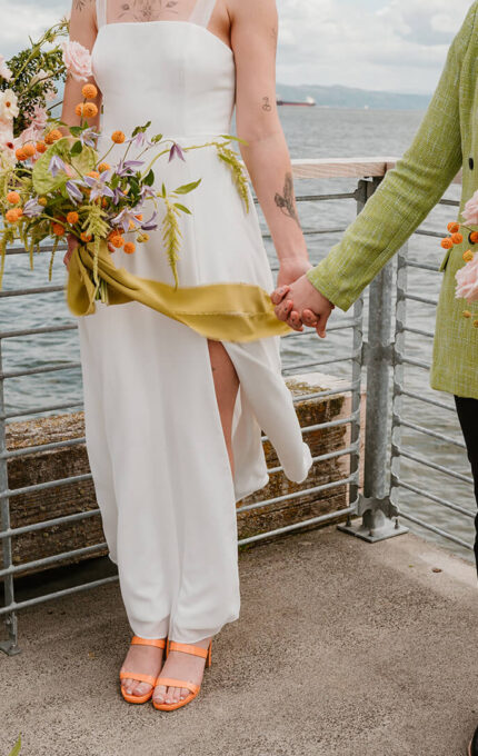 Couple holding hands with colourful flower bouquets on scenic waterfront terrace, ideal for romantic getaways or weddings.