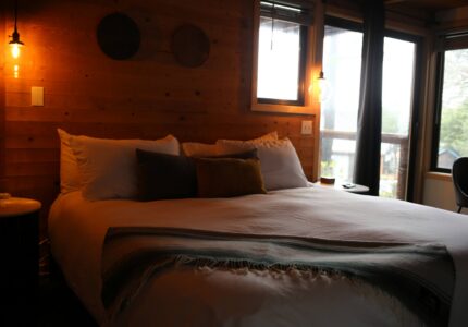 Cosy hotel room with king bed, rustic wood walls, warm lighting, and large windows offering serene nature views.