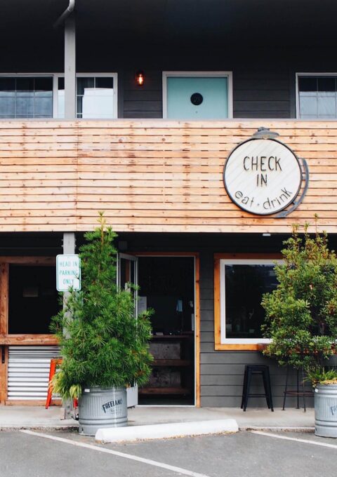 Check In, Eat, Drink sign at the entrance of Ashore Hotel in Seaside, Oregon, with wood-paneled facade and potted plants.