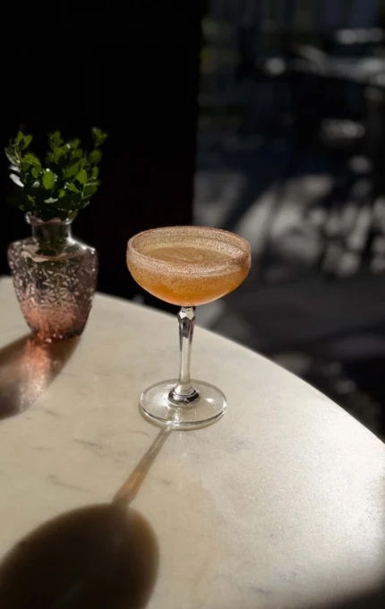 Sparkling amber cocktail in a coupe glass on a sunlit marble table with decorative vase and greenery — relaxing hotel bar ambience